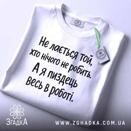 Футболка для тих хто працює. Біла футболка з чорним принтом, складена на столі з верхнього ракурсу. Фон світло-сірий, на футболці видно текст: 'Не лається той, хто нічого не робить. А я піздець весь в роботі.'