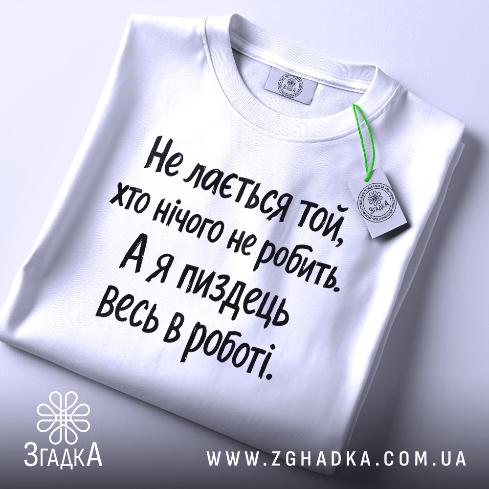 Футболка для тих хто працює. Біла футболка з чорним принтом, складена на столі з верхнього ракурсу. Фон світло-сірий, на футболці видно текст: 'Не лається той, хто нічого не робить. А я піздець весь в роботі.'