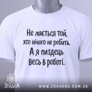 Футболка для тих хто працює. Біла футболка з чорним принтом, складена на столі з верхнього ракурсу. Фон світло-сірий, на футболці видно текст: 'Не лається той, хто нічого не робить. А я піздець весь в роботі.'