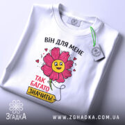 Футболка для закоханих сердець, білого кольору, складена на поверхні, фон світло-сірий, принт з квіткою та написом "ВІН ДЛЯ МЕНЕ ТАК БАГАТО ЗНАЧИТЬ!", на футболці є етикетка з зеленим шнурком.