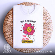Футболка для закоханих сердець, білого кольору, складена на плетеній кошику, фон світлий, принт з квіткою та написом "ВІН ДЛЯ МЕНЕ ТАК БАГАТО ЗНАЧИТЬ!".