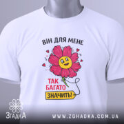 Футболка для закоханих сердець, білого кольору, складена на поверхні, фон світло-сірий, принт з квіткою та написом "ВІН ДЛЯ МЕНЕ ТАК БАГАТО ЗНАЧИТЬ!", на футболці є етикетка з зеленим шнурком.