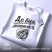 Футболка до біса досконалість, біла, складена на столі, фон світло-сірий, принт чорний з написом 'До біса досконалість' і абстрактним малюнком, на комірці зелена етикетка.