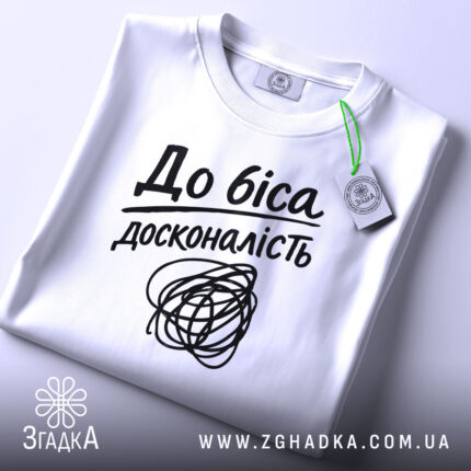 Футболка до біса досконалість, біла, складена на столі, фон світло-сірий, принт чорний з написом 'До біса досконалість' і абстрактним малюнком, на комірці зелена етикетка.