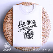 Футболка до біса досконалість, біла, складена на плетеній кошику, фон світло-сірий, принт чорний з написом 'До біса досконалість' і абстрактним малюнком.