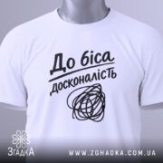 Футболка до біса досконалість, біла, складена на столі, фон світло-сірий, принт чорний з написом 'До біса досконалість' і абстрактним малюнком, на комірці зелена етикетка.