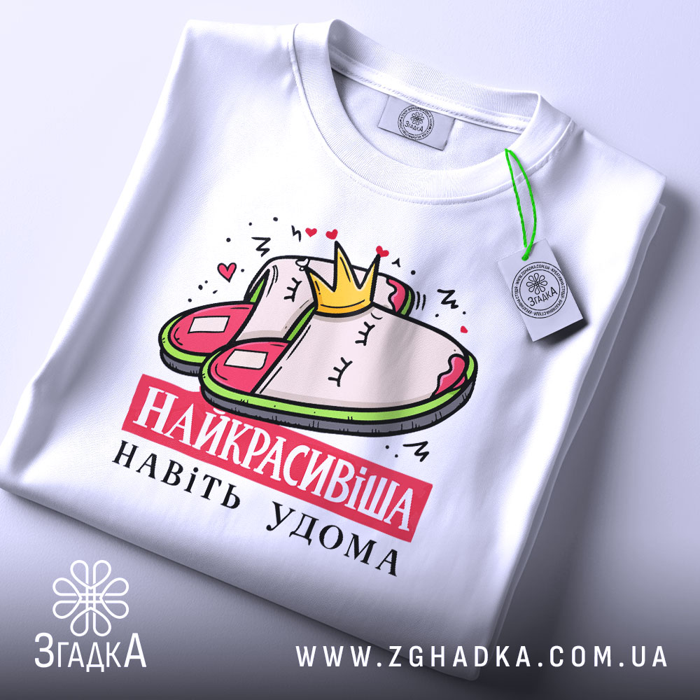 Футболка домашня королева, біла, складена на столі, фон світло-сірий, принт з малюнком домашніх капців і корони, текст "НАЙКРАСИВІША НАВІТЬ УДОМА" червоним кольором, на футболці є етикетка.