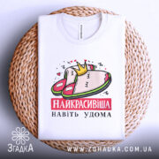 Футболка домашня королева, біла, складена на плетеному кошику, фон світло-сірий, принт з малюнком домашніх капців і корони, текст "НАЙКРАСИВІША НАВІТЬ УДОМА" червоним кольором.