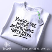 Футболка досягати через ліжко, біла, складена на поверхні, з видимим етикеткою, фон світлий, принт чорний з текстом, що говорить про бажання досягти чогось через ліжко.
