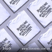 Футболка досягати через ліжко, біла, зображена з фронтального ракурсу на манекені, фон світлий, принт чорний з текстом, що говорить про бажання досягти чогось через ліжко.