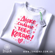 Футболка дуже сильно кохаю, білого кольору, складена на поверхні, фон світло-сірий, принт червоного кольору з написом 'Дуже сильно тебе кохаю!' та етикетка з зеленим шнурком.