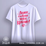 Футболка дуже сильно кохаю, білого кольору, підвішена на вішалці, фон світло-сірий, принт червоного кольору з написом 'Дуже сильно тебе кохаю!' та декоративними елементами.