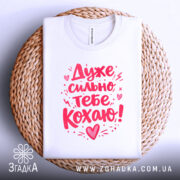 Футболка дуже сильно кохаю, білого кольору, складена на плетеній кошику, фон світло-сірий, принт червоного кольору з написом 'Дуже сильно тебе кохаю!'.