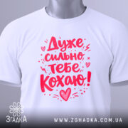 Футболка дуже сильно кохаю, білого кольору, складена на поверхні, фон світло-сірий, принт червоного кольору з написом 'Дуже сильно тебе кохаю!' та етикетка з зеленим шнурком.