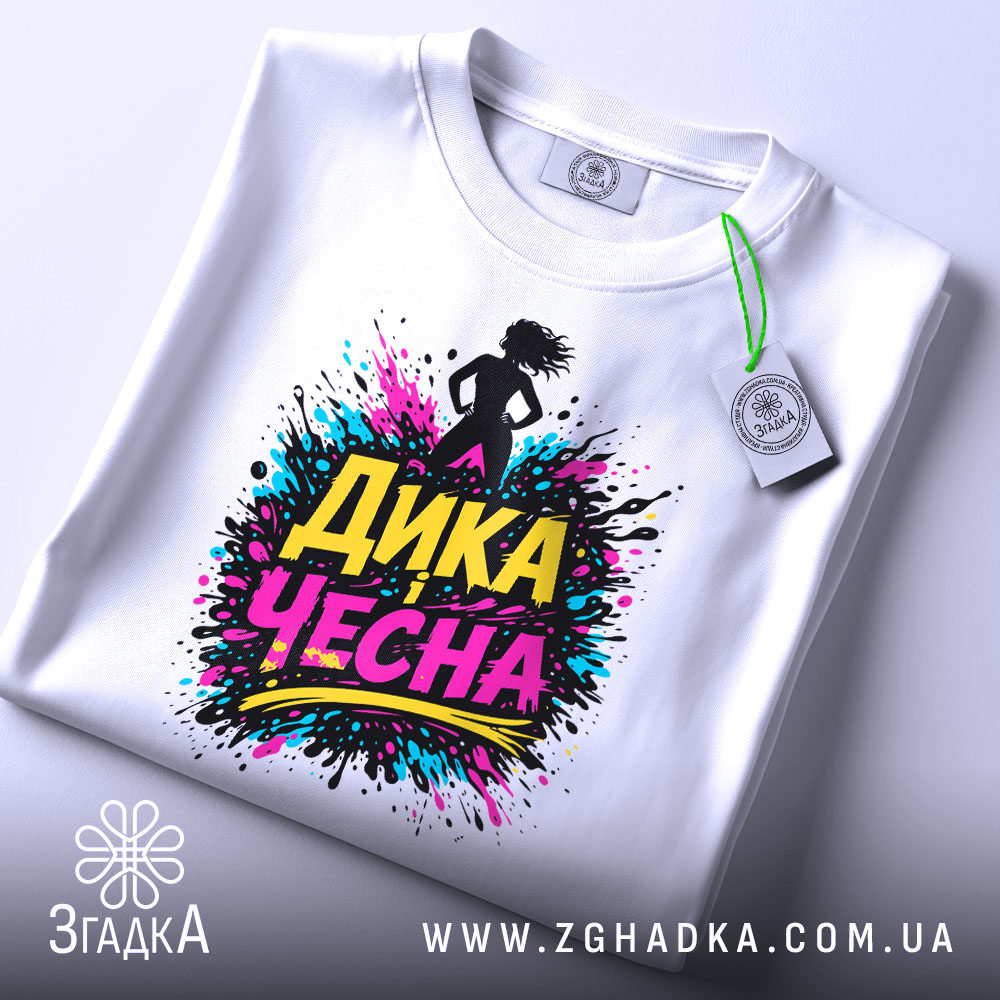 Футболка дика чесна, біла, складена на плоскій поверхні, фон світлий, принт з написом 'ДИКА ЧЕСНА' в яскравих кольорах, прикріплена етикетка з зеленим шнурком.