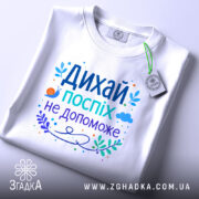 Футболка дихай спокійно, біла, зображена зверху, складена на поверхні, фон світлий, принт з текстом "Дихай, поспіх не допоможе" в яскравих кольорах, з елементами рослинності та малюнком равлика, на футболці є етикетка.