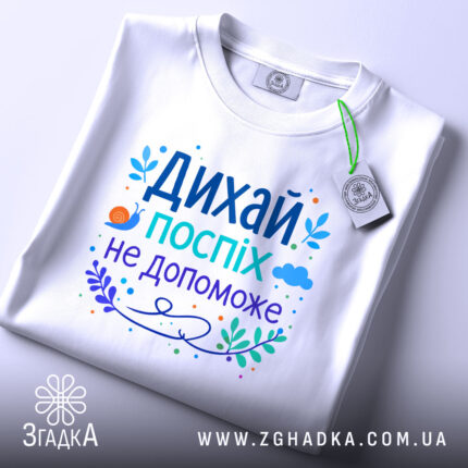 Футболка дихай спокійно, біла, зображена зверху, складена на поверхні, фон світлий, принт з текстом "Дихай, поспіх не допоможе" в яскравих кольорах, з елементами рослинності та малюнком равлика, на футболці є етикетка.