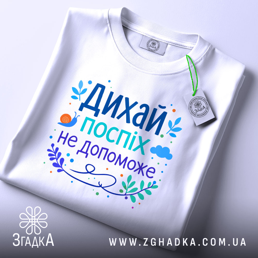 Футболка дихай спокійно, біла, зображена зверху, складена на поверхні, фон світлий, принт з текстом "Дихай, поспіх не допоможе" в яскравих кольорах, з елементами рослинності та малюнком равлика, на футболці є етикетка.