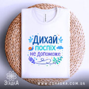 Футболка дихай спокійно, біла, зображена зверху, складена на плетеній основі, фон світлий, принт містить текст "Дихай, поспіх не допоможе" в яскравих кольорах, з елементами рослинності та малюнком равлика.