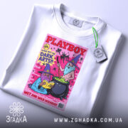 Футболка дикі чарівники, біла, зображена зверху, на ній є етикетка з зеленим шнурком, фон світлий, принт з двома чарівниками, один читає книгу, інший варить зілля, яскраві кольори на рожевому фоні.