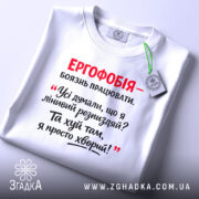 Футболка ергофобія гумор. Біла футболка, складена на поверхні, з принтом червоного та чорного кольору. На фоні видно етикетку з зеленим шнурком.