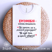 Футболка ергофобія гумор. Біла футболка, складена на плетеній кошику, з принтом червоного та чорного кольору. Фон світлий, акцент на тексті жарту.