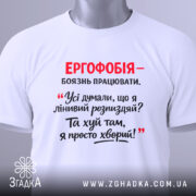 Футболка ергофобія гумор. Біла футболка, складена на поверхні, з принтом червоного та чорного кольору. На фоні видно етикетку з зеленим шнурком.