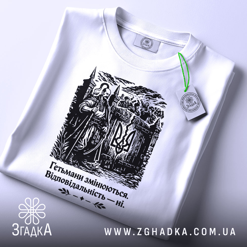 Футболка гетьмани змінюються. Біла футболка з чорним принтом, зображення гетьмана на фоні замку, ракурс зверху, видно етикетку з зеленим шнурком, принт чітко виділяється на фоні.