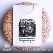 Футболка гетьмани змінюються. Біла футболка з чорним принтом, зображення гетьмана на фоні замку, ракурс зверху, футболка лежить на плетеній основі, принт чітко видимий.