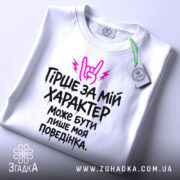 Футболка гірше ніж характер. Біла футболка, складена на столі, з принтом чорного кольору та рожевими елементами. На фоні світло-сірого кольору, видно етикетку з зеленим шнурком.