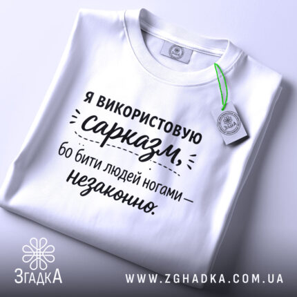 Футболка гострий сарказм, біла, складена на поверхні, фон світло-сірий, принт чорного кольору з текстом про сарказм, з зеленим ярликом.
