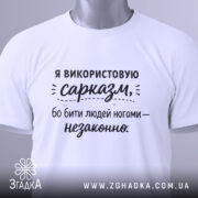 Футболка гострий сарказм, біла, складена на поверхні, фон світло-сірий, принт чорного кольору з текстом про сарказм, з зеленим ярликом.