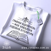 Футболка графіки відключень, білого кольору, зображена на столі з ракурсу зверху, фон світло-сірий, принт чорного кольору з малюнком електричної опори та відкритої книги, на футболці є етикетка з зеленим шнурком.