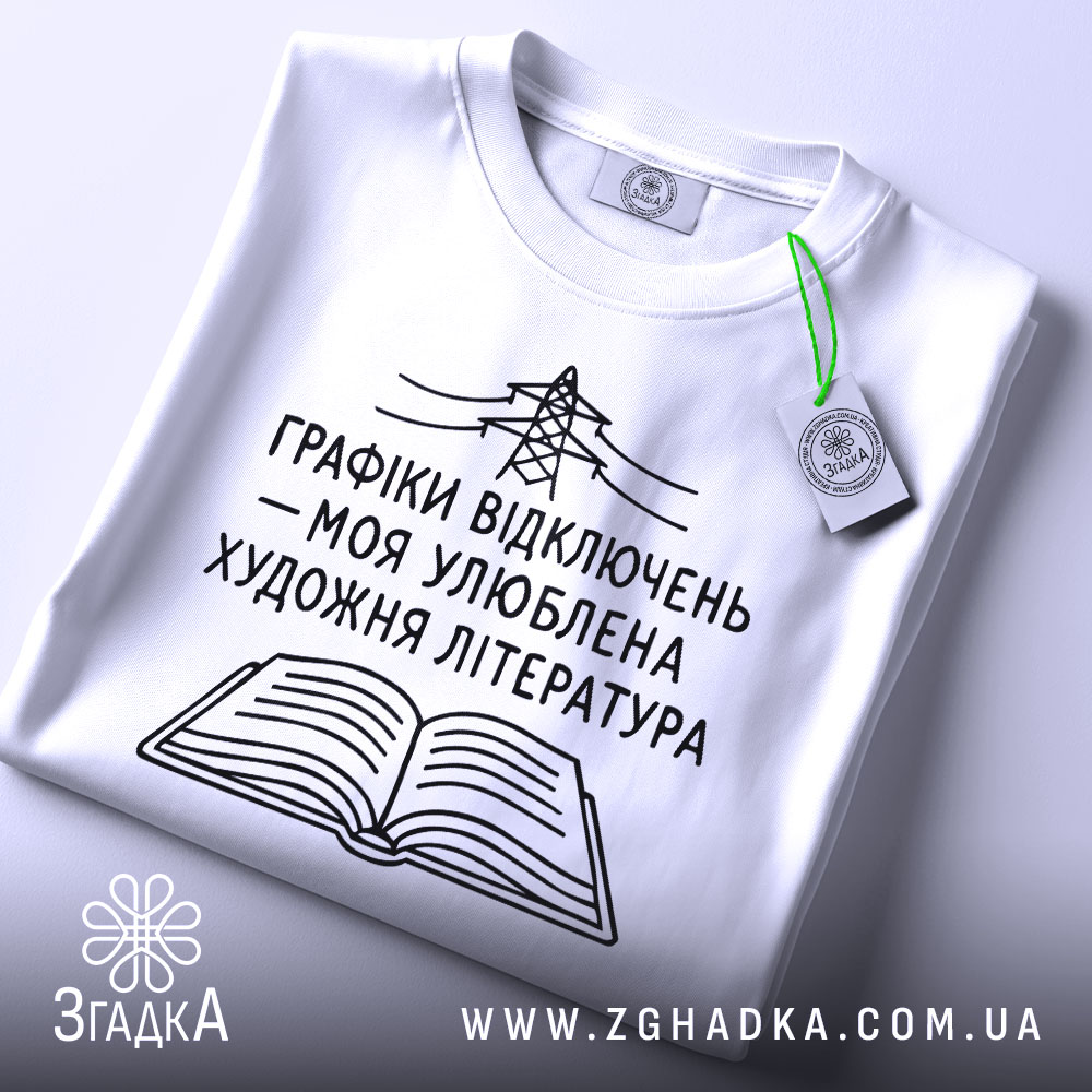 Футболка графіки відключень, білого кольору, зображена на столі з ракурсу зверху, фон світло-сірий, принт чорного кольору з малюнком електричної опори та відкритої книги, на футболці є етикетка з зеленим шнурком.