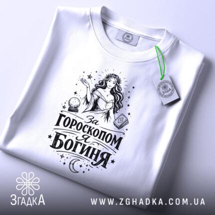 Футболка я богиня, біла, складена на поверхні, з видимим етикеткою, фон світло-сірий, принт чорного кольору з жінкою та астрологічними елементами.