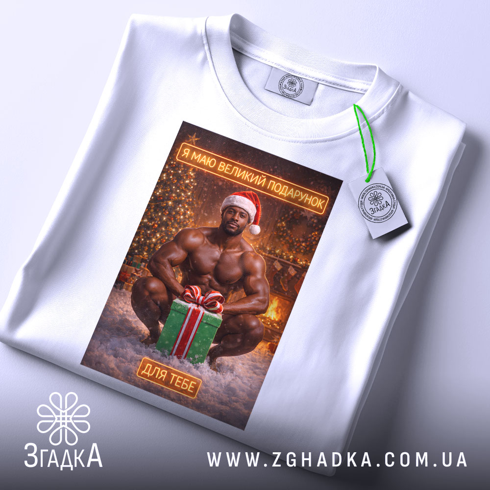 Футболка я маю подарунок, біла, з ракурсу зверху, на фоні світлого кольору, принт з зображенням чоловіка в червоній шапці Санти, який сидить на снігу з подарунком, з етикеткою на рукаві.