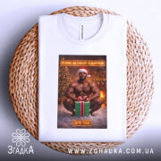 Футболка я маю подарунок, біла, з ракурсу зверху, на фоні плетеного кошика, принт з зображенням чоловіка в червоній шапці Санти, який сидить на снігу з подарунком.