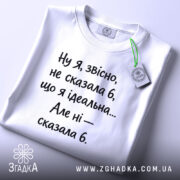 Футболка я не ідеальна. Біла футболка, складена на столі, з коротким рукавом. Принт чорного кольору з текстом, прикріплена бірка зеленого кольору.