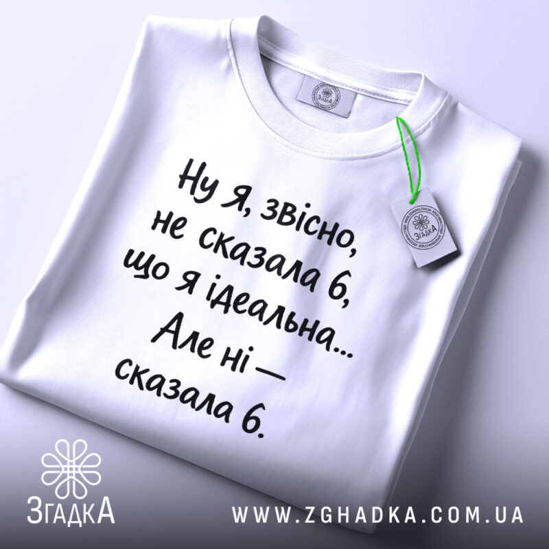 Футболка я не ідеальна. Біла футболка, складена на столі, з коротким рукавом. Принт чорного кольору з текстом, прикріплена бірка зеленого кольору.
