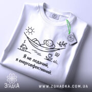 Футболка я не ледачий, біла, зображена на столі, фон світло-сірий, принт з малюнком людини в гамаку, текст чорного кольору, на комірці є етикетка.