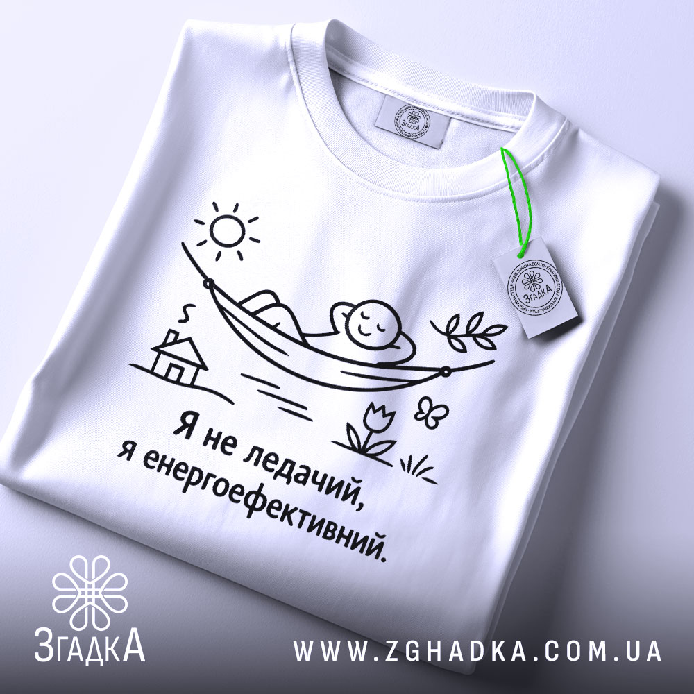 Футболка я не ледачий, біла, зображена на столі, фон світло-сірий, принт з малюнком людини в гамаку, текст чорного кольору, на комірці є етикетка.