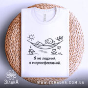 Футболка я не ледачий, біла, зображена на плетеному кошику, фон світло-сірий, принт з малюнком людини в гамаку, текст чорного кольору.