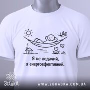 Футболка я не ледачий, біла, зображена на столі, фон світло-сірий, принт з малюнком людини в гамаку, текст чорного кольору, на комірці є етикетка.
