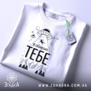 Футболка я обираю тебе. Біла футболка, складена на столі, з видимим етикеткою. На фоні світло-сірий градієнт. Принт з НЛО, горами та написом 'Я обираю ТЕБЕ' чорного кольору.