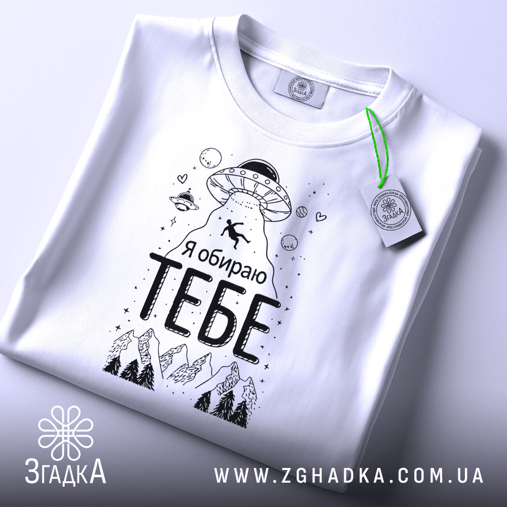 Футболка я обираю тебе. Біла футболка, складена на столі, з видимим етикеткою. На фоні світло-сірий градієнт. Принт з НЛО, горами та написом 'Я обираю ТЕБЕ' чорного кольору.