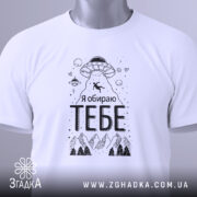 Футболка я обираю тебе. Біла футболка, складена на столі, з видимим етикеткою. На фоні світло-сірий градієнт. Принт з НЛО, горами та написом 'Я обираю ТЕБЕ' чорного кольору.