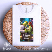 Футболка я обираю тебе, біла, складена на плетеній основі, з принтом, що зображує Шрека та Фіону, на фоні казкового лісу.