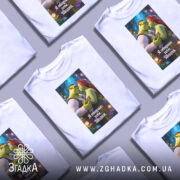 Футболка я обираю тебе, біла, з принтом, що зображує персонажів Шрека та Фіони, на фоні казкового лісу з кольоровими вогниками.