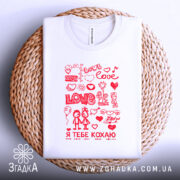 Футболка я тебе кохаю, біла, складена на плетеній кошику, фон світло-сірий, принт червоний з малюнками сердець, квітів та двох персонажів, які тримаються за руки.