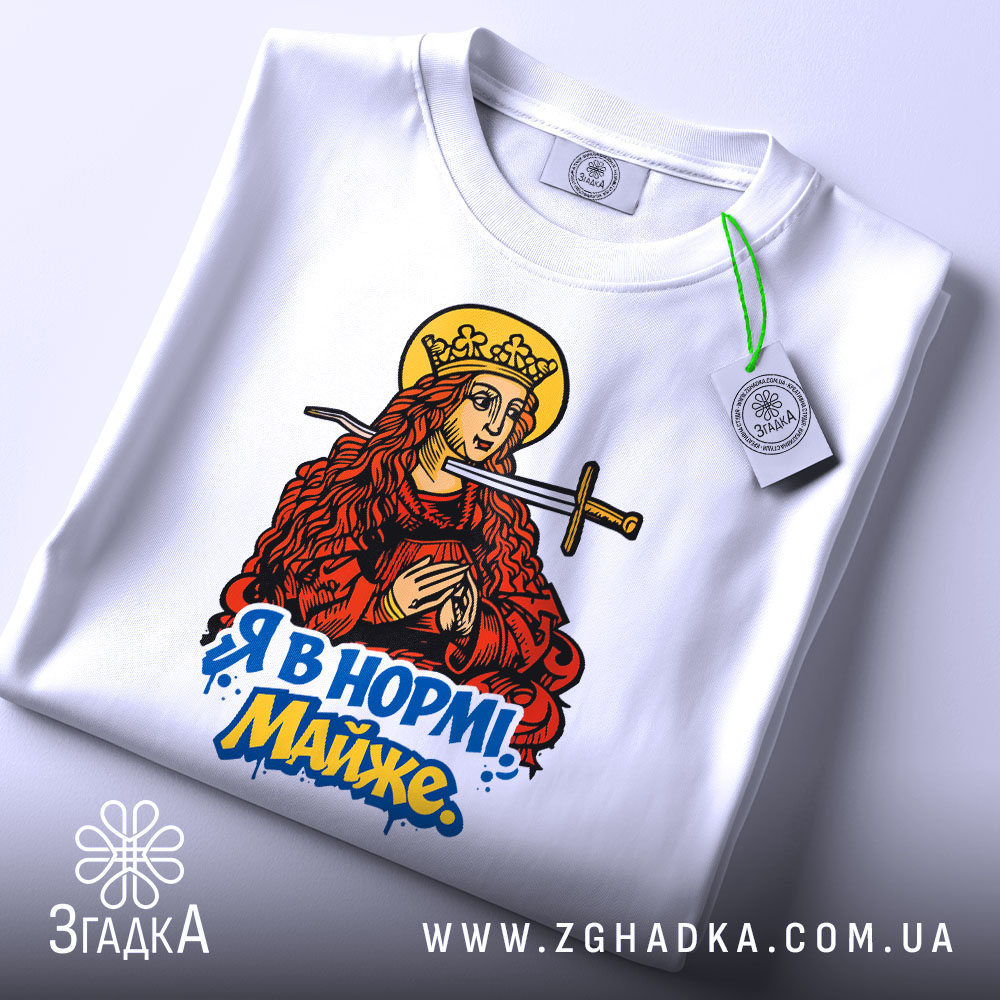 Футболка я в нормі майже білого кольору, складена на поверхні, фон світло-сірий, принт з червоно-золотистою жінкою з мечем та написом "Я в нормі! Майже" у яскравих кольорах, з зеленим ярликом.
